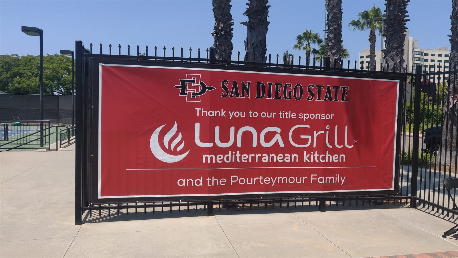 Red sponsor banner for San Diego State and Luna Grill mounted along a tennis court fence